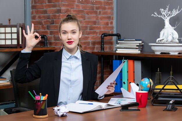 Draufsicht einer jungen Dame, die an einem Tisch sitzt und das Dokument hält, das im Büro eine Brillengeste macht