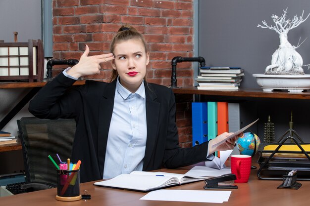 Draufsicht einer jungen Frau, die an einem Tisch sitzt und ein Dokument hält, das im Büro eine Waffengeste macht