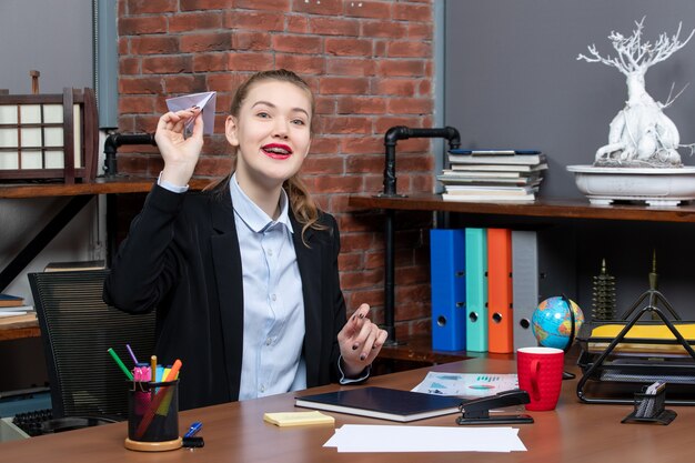 Draufsicht einer jungen, glücklich lächelnden Büroangestellten, die an ihrem Schreibtisch sitzt und Papierflieger spielt