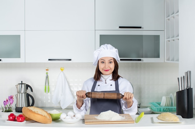 Draufsicht einer Köchin in Uniform, die hinter dem Tisch steht, mit Schneidebrett-Lebensmitteln, die Nudelholz in der weißen Küche halten