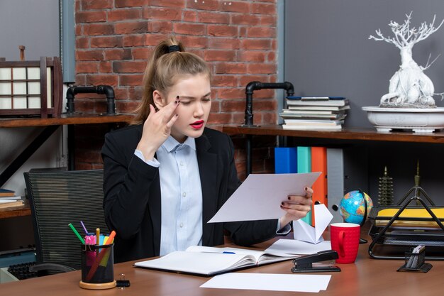 Draufsicht einer konzentrierten jungen Frau, die an einem Tisch sitzt und ein Dokument hält, das etwas sorgfältig im Büro liest