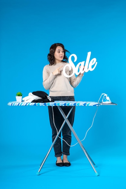 Draufsicht einer konzentrierten jungen Frau, die hinter dem Bügelbrett steht und das Verkaufssymbol auf blauem Hintergrund zeigt