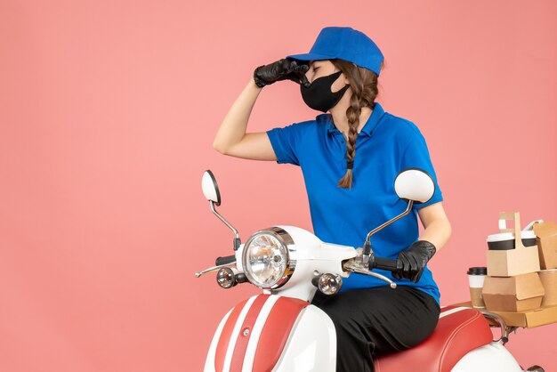 Draufsicht einer Kurierfrau mit medizinischer Maske und Handschuhen, die auf einem Roller sitzt und Bestellungen liefert, die eine schlechte Geruchsgeste auf pastellfarbenem Pfirsichhintergrund machen