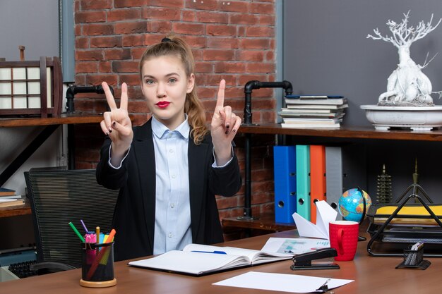 Draufsicht einer lächelnden jungen Frau, die an einem Tisch sitzt und nach oben zeigt und im Büro eine Siegesgeste macht