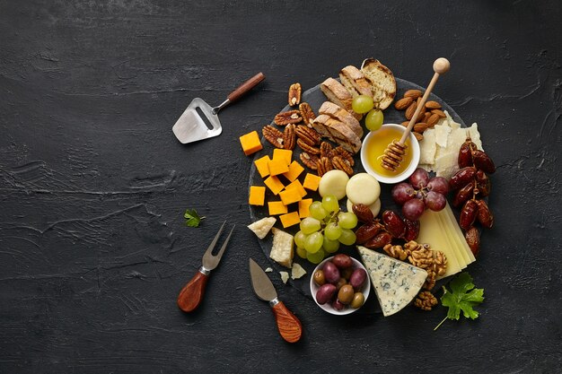 Draufsicht einer leckeren Käseplatte mit Obst, Trauben, Nüssen und Honig auf einer kreisförmigen Küchenplatte auf schwarzem Steinhintergrund, Draufsicht, Kopierraum. Gourmetessen und trinken.