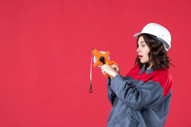 Draufsicht einer neugierigen Architektin in Uniform mit Schutzhelm, die Maßband auf isoliertem rotem Hintergrund hält