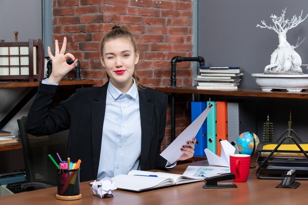 Draufsicht einer selbstbewussten jungen Dame, die an einem Tisch sitzt und das Dokument hält, das im Büro eine Brillengeste macht