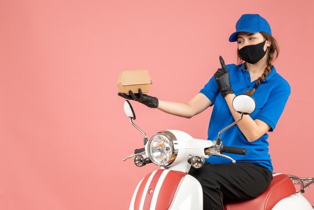 Draufsicht einer selbstbewussten Lieferperson mit medizinischer Maske und Handschuhen, die auf einem Roller sitzt und Bestellungen auf pastellfarbenem Pfirsichhintergrund liefert