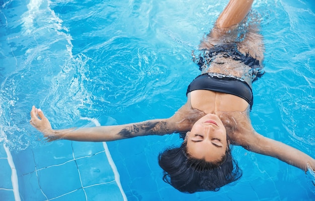 Draufsicht einer sinnlich entspannten Frau im schwarzen Bikini mit geschlossenen Augen, die im Pool auf dem Rücken schwimmt Attraktives sexy weibliches Modell, das kühles Wasser im Schwimmbad im Sommertourismus und Sommerzeitkonzept genießt