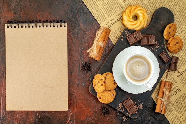 Draufsicht einer Tasse Kaffee auf einem Holzbrett auf einem alten Zeitungsplätzchen-Zimt-Limonen-Schokoriegel-Notizbuch auf dunkler Oberfläche