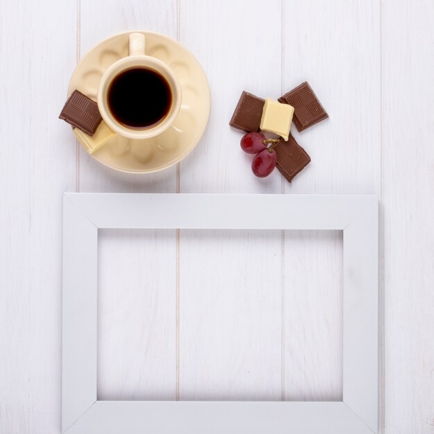 Draufsicht einer Tasse Kaffee mit weißer und dunkler Schokolade und einem leeren Bilderrahmen auf weißem hölzernem Hintergrund mit Kopienraum