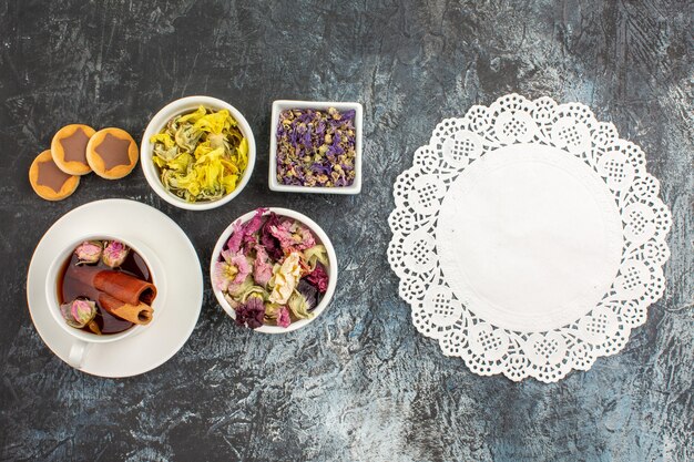 Draufsicht einer Tasse Kräutertee mit Keksen und getrockneten Blumen und weißer Spitze auf grauem Hintergrund