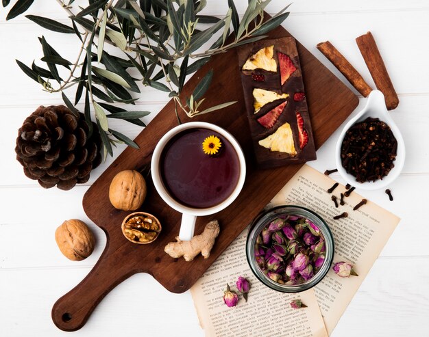 Draufsicht einer Tasse Tee mit Schokoriegel mit getrockneten Früchten und ganzen Walnüssen auf Holzschneidebrett, trockenen Rosenknospen in einem Glas und Gewürznelke auf Weiß