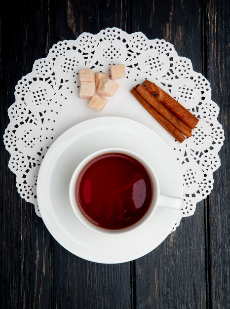 Draufsicht einer Tasse Tee mit Zimtstangen und braunen Zuckerwürfeln auf Spitzenpapierserviette auf dunklem hölzernem Hintergrund