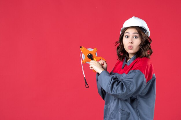 Draufsicht einer überraschten Architektin in Uniform mit Schutzhelm, der Maßband auf isoliertem rotem Hintergrund hält