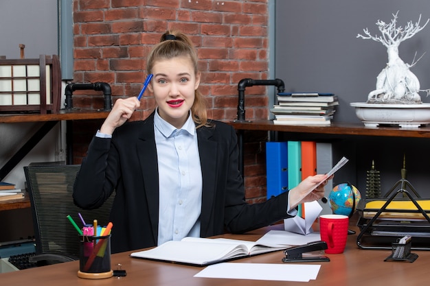 Kostenloses Foto draufsicht einer verwirrten frau, die an einem tisch sitzt und einen blauen farbstift des dokuments im büro hält