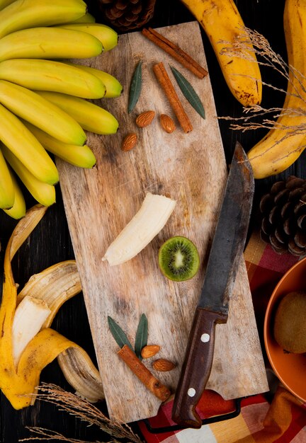 Draufsicht eines Bündels von Bananenfrüchten und geschnittener Banane auf einem hölzernen Schneidebrett mit einem Messer und Zimtstangen auf rustikalem