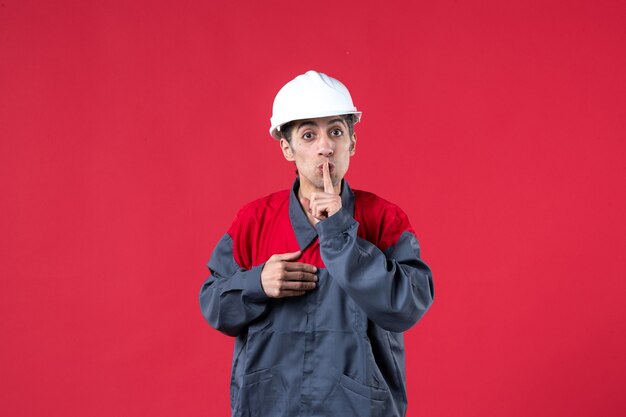 Draufsicht eines ernsten jungen Baumeisters in Uniform mit Schutzhelm, der auf isolierter roter Wand Stillegeste macht