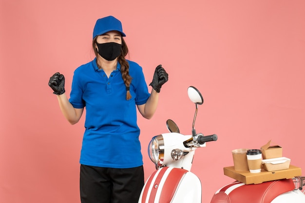 Draufsicht eines glücklichen Kuriermädchens, das medizinische Maskenhandschuhe trägt, die neben dem Motorrad mit Kaffeekuchen darauf auf pastellfarbenem Hintergrund in Pfirsichfarbe steht