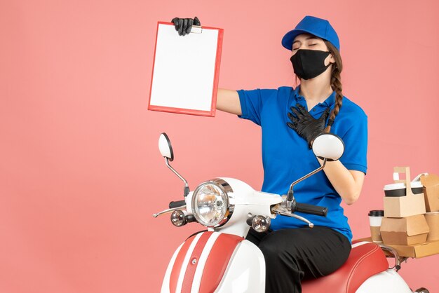 Draufsicht eines hoffnungsvollen Kuriermädchens mit medizinischer Maske und Handschuhen, das auf einem Roller sitzt und ein leeres Papierblatt hält, das Bestellungen auf pastellfarbenem Pfirsichhintergrund liefert delivering