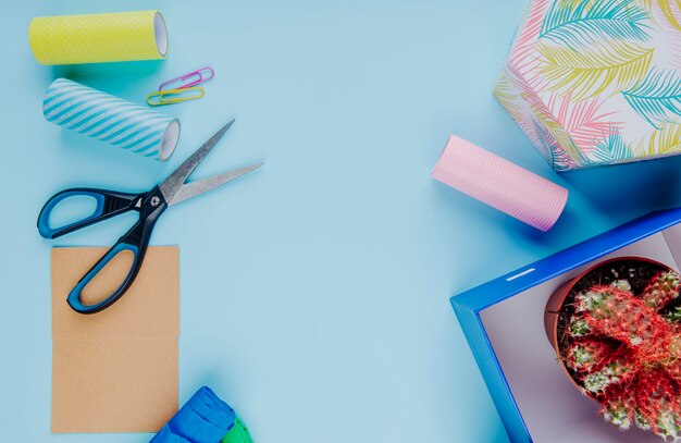 Draufsicht eines kaktus in einem blumentopf in einer geschenkbox des kartons mit bissigen büroklammern der schere und rollen des klebebands auf blauem hintergrund