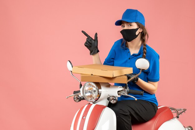 Draufsicht eines konzentrierten weiblichen Kuriers mit medizinischer Maske und Handschuhen, der auf einem Roller sitzt und Bestellungen auf pastellfarbenem Pfirsichhintergrund liefert