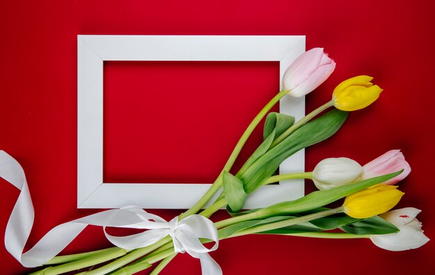 Draufsicht eines leeren Bilderrahmens mit einem Strauß bunter Tulpenblumen auf rotem Hintergrund mit Kopienraum