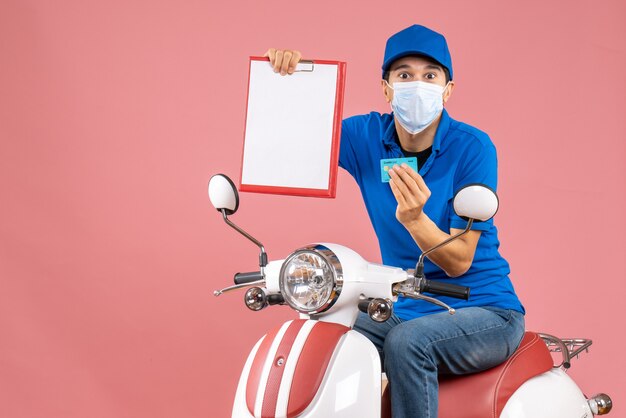 Draufsicht eines männlichen Lieferers in Maske mit Hut, der auf einem Roller sitzt und ein Dokument und eine Bankkarte auf pastellfarbenem Pfirsichhintergrund zeigt