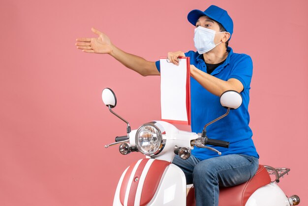 Draufsicht eines männlichen Lieferers in Maske mit Hut, der auf einem Roller sitzt und ein Dokument zeigt, das etwas auf pastellfarbenem Pfirsichhintergrund fragt