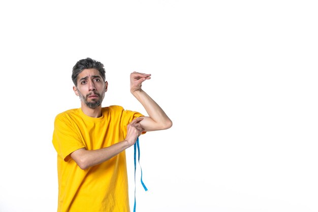 Draufsicht eines nervösen jungen Mannes in gelbem Hemd, der ein Messgerät hält und seine Muskeln auf weißer Oberfläche misst