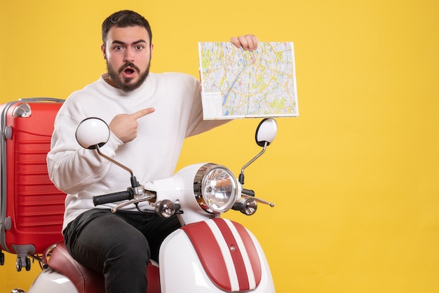 Draufsicht eines neugierigen jungen Mannes, der auf einem Motorrad mit Koffer darauf sitzt und Karte auf isoliertem gelbem Hintergrund hält