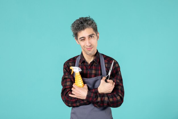 Draufsicht eines selbstbewussten Friseurs, der eine graue Schürze trägt und eine Schere-Sprühflasche auf pastellblauem Hintergrund hält