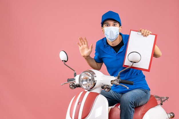 Draufsicht eines überraschten männlichen Lieferers in Maske mit Hut, der auf einem Roller sitzt und ein Dokument auf Pastellpfirsich zeigt