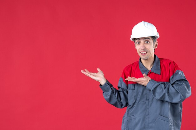 Draufsicht eines verwirrten jungen Arbeiters in Uniform mit Schutzhelm auf isolierter roter Wand