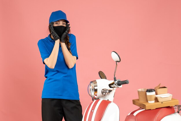 Draufsicht eines verwirrten Kuriermädchens, das medizinische Maskenhandschuhe trägt, die neben dem Motorrad mit Kaffeekuchen darauf auf pastellfarbenem Pfirsichhintergrund steht standing