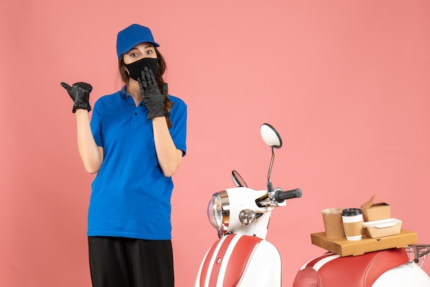 Draufsicht eines verwirrten Kuriermädchens in medizinischer Maske, das neben dem Motorrad mit Kaffeekuchen auf pastellfarbenem Hintergrund steht