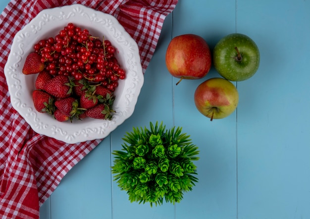 Draufsicht Erdbeeren mit roten Johannisbeeren auf einem Teller mit Äpfeln und einem roten Küchentuch auf einem hellblauen Hintergrund