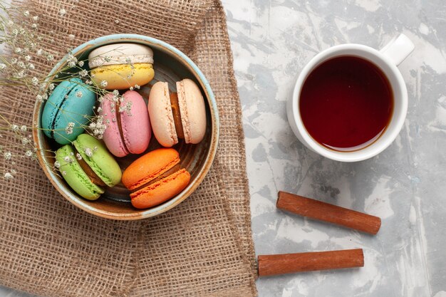 Draufsicht farbige französische Macarons köstliche kleine Kuchen mit Tasse Tee auf weißem Schreibtisch