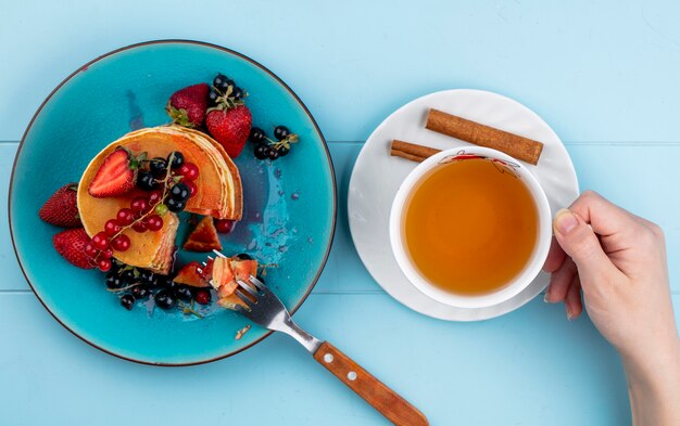 Draufsicht Frau trinkt Tasse Tee mit Pfannkuchen mit Erdbeeren roten und schwarzen Johannisbeeren auf einem blauen Tisch