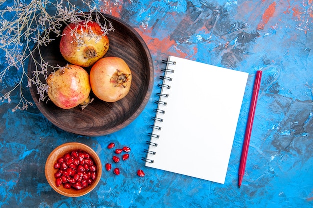 Draufsicht frische granatäpfel in holzschale granatapfelkerne in einer schüssel ein roter notizbuchstift auf blauem hintergrund