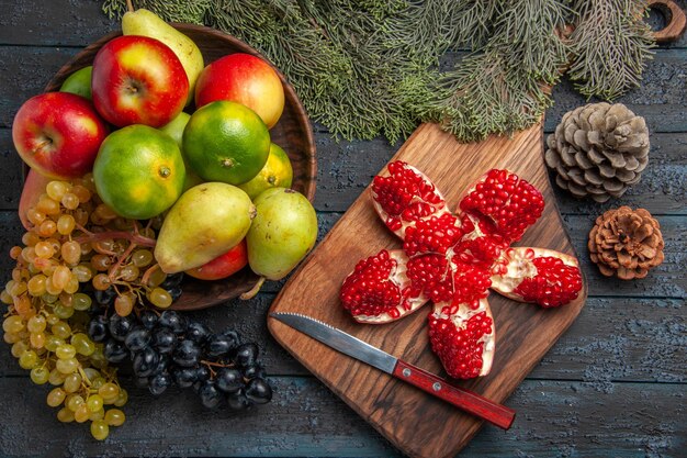 Draufsicht Früchte und Zweige weiße und schwarze Trauben Limetten Birnen Äpfel in Schüssel neben Granatapfelmesser auf Küchenbrett und Fichtenzweigen mit Zapfen auf dunklem Tisch