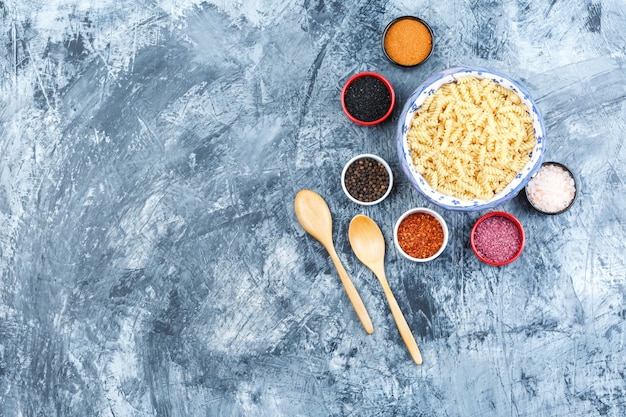 Draufsicht-Fusilli-Nudeln in der Schüssel mit Gewürzen, Holzlöffel auf grauem Gipshintergrund. horizontal