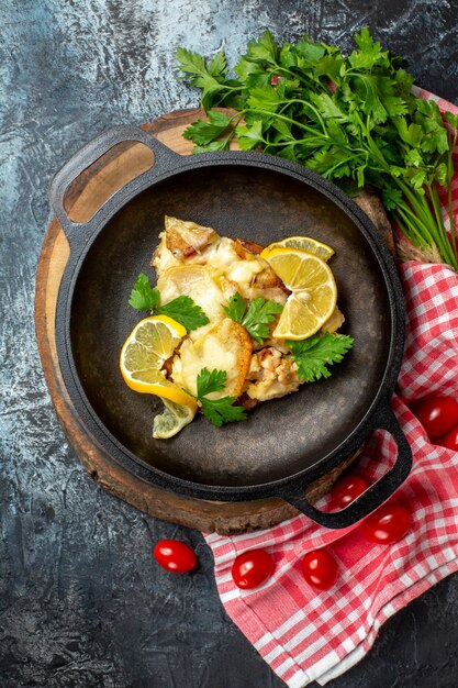 Draufsicht gebratener Fisch in Pfanne auf Holzbrett Kirschtomaten Petersilie auf rot-weiß karierter Tischdecke auf grauem Hintergrund