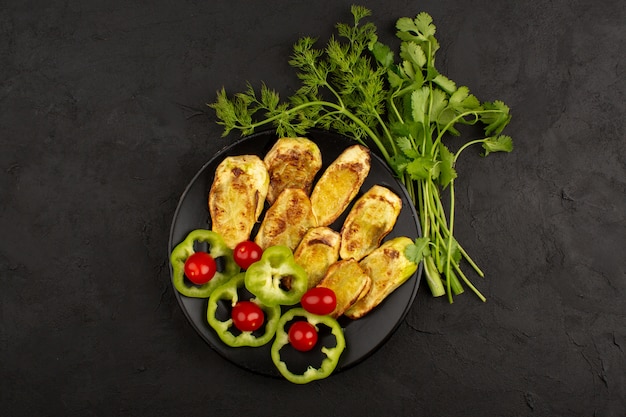 Draufsicht gekochte Aubergine in schwarzer Platte zusammen mit geschnittenem grünem Paprika und roten Kirschtomaten auf der Dunkelheit