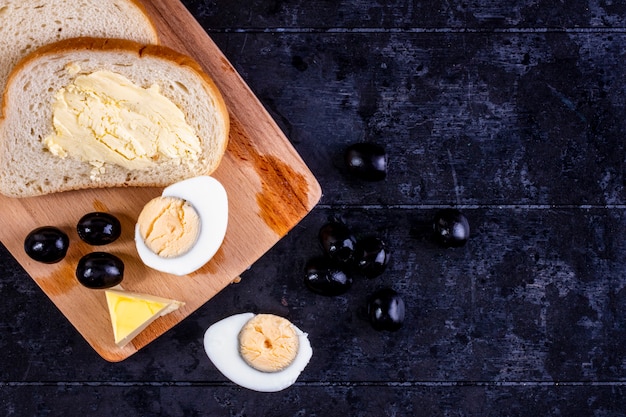 Kostenloses Foto draufsicht gekochtes ei an bord mit oliven und scheiben brot und butter auf schwarz