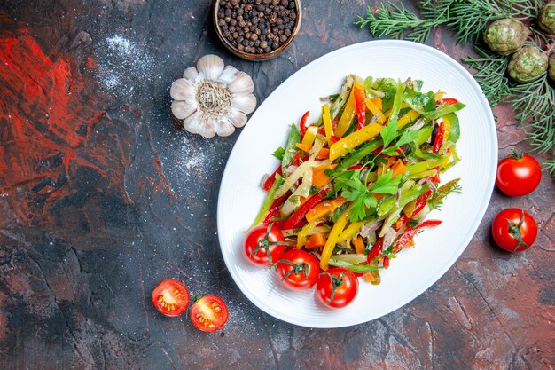 Draufsicht Gemüsesalat auf ovalem Teller Knoblauchkirschtomaten schwarzer Pfeffer auf dunkelrotem Tisch