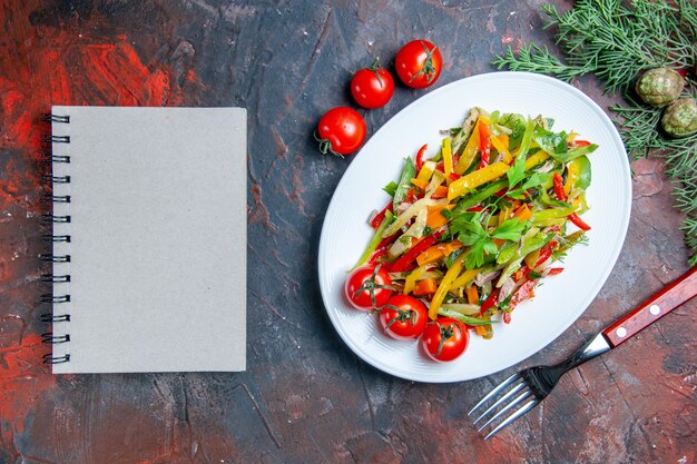 Draufsicht Gemüsesalat auf ovalem Tellergabel-Kirschtomaten-Notizbuch auf dunkelrotem Tisch