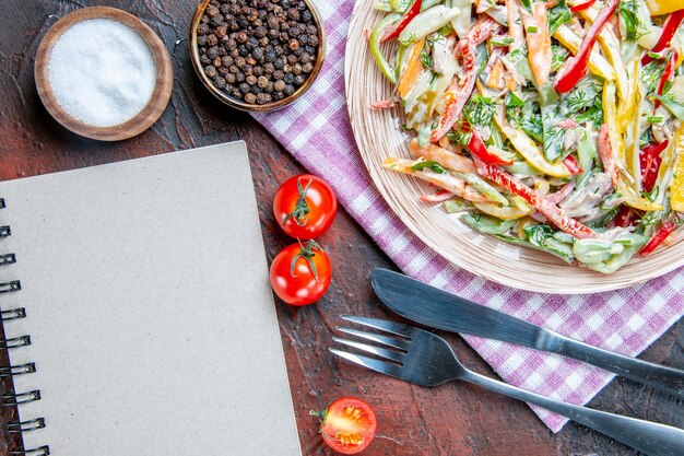 Draufsicht Gemüsesalat auf Teller auf Tischdecke Gabel und Messersalz und Tomaten mit schwarzem Pfeffer ein Notizbuch auf dunkelrotem Tisch