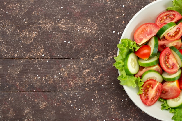 Draufsicht geschnittene Tomaten mit Gurken in weißem Teller mit grünem Salat auf braunem, frischem Gemüsesalat zum Mittagessen