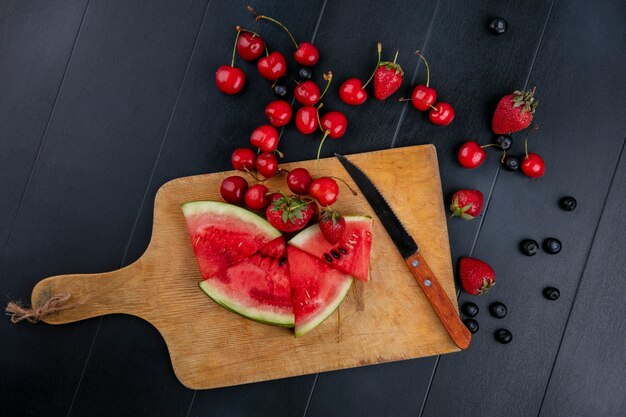 Draufsicht geschnittene Wassermelone auf einem Brett mit Erdbeeren und Kirschen mit einem Messer auf einem schwarzen Hintergrund